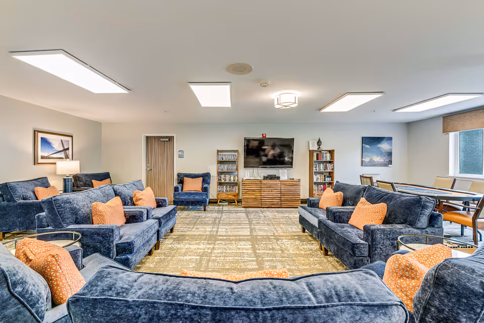 A spacious senior living common area with multiple blue velvet sofas and armchairs arranged around a large patterned carpet. The sofas have orange throw pillows. A flat-screen TV is mounted on the wall above a wooden media console, flanked by bookshelves filled with books and DVDs. There are framed pictures on the walls and a window with a brown blind letting in natural light. A table with chairs is visible near the window.