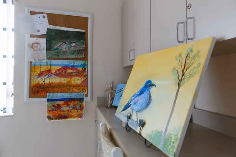 Art workspace with several paintings displayed on a countertop and pinned to a corkboard in a light interior room.