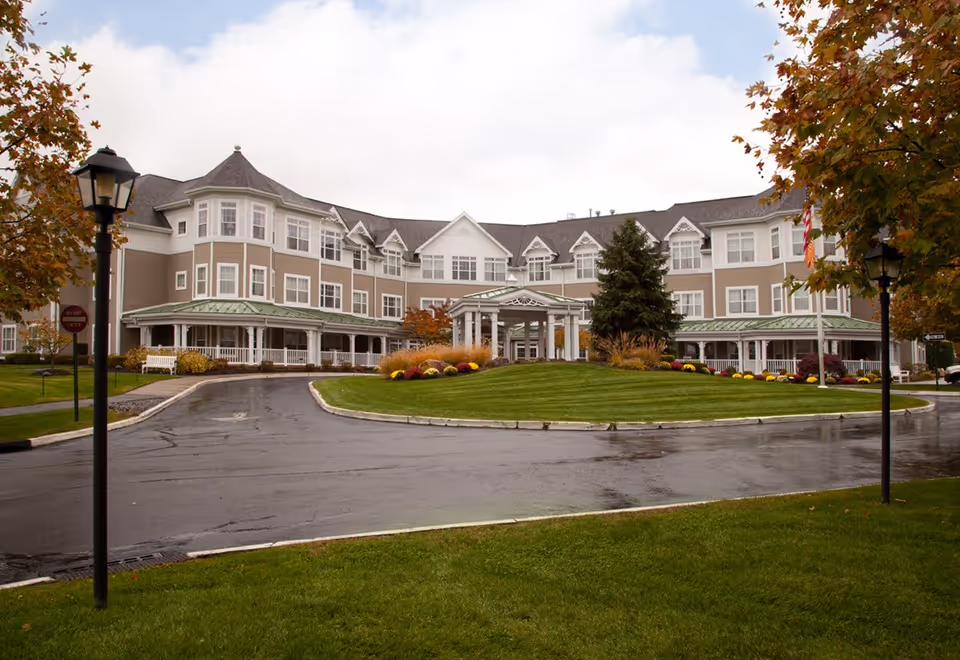 Exterior view of a large, multi-story senior living facility with beige siding and white trim. The building has multiple windows, a covered entrance with white columns, and a circular driveway surrounded by well-maintained green lawns and colorful flower beds. Trees with autumn foliage and lampposts line the driveway.