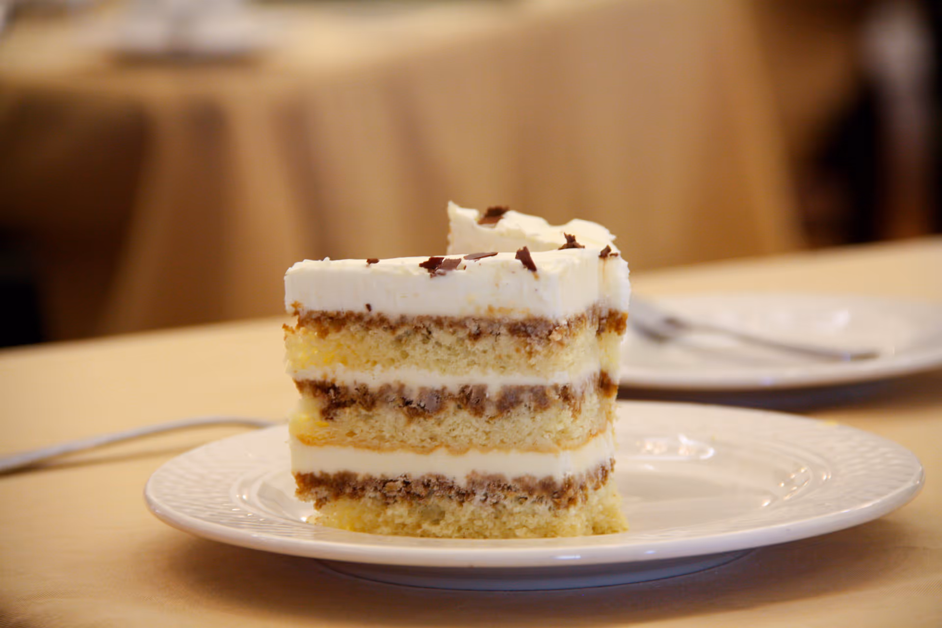 A slice of layered cake with white frosting and chocolate shavings on top, served on a white plate with a blurred background of a table and another plate with a fork.