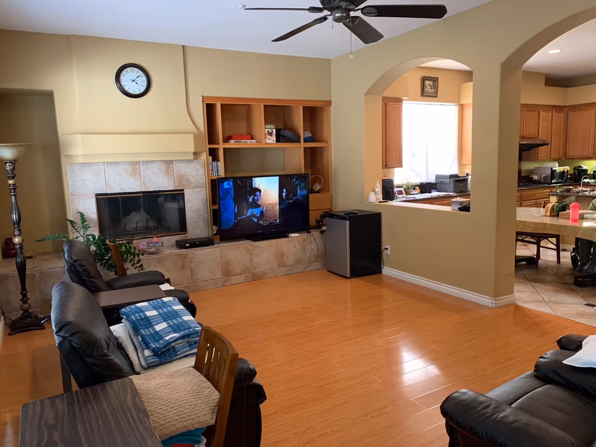Open living room with a fireplace and clock, built-in shelving holding a TV, leather recliners, and an archway opening into the kitchen.