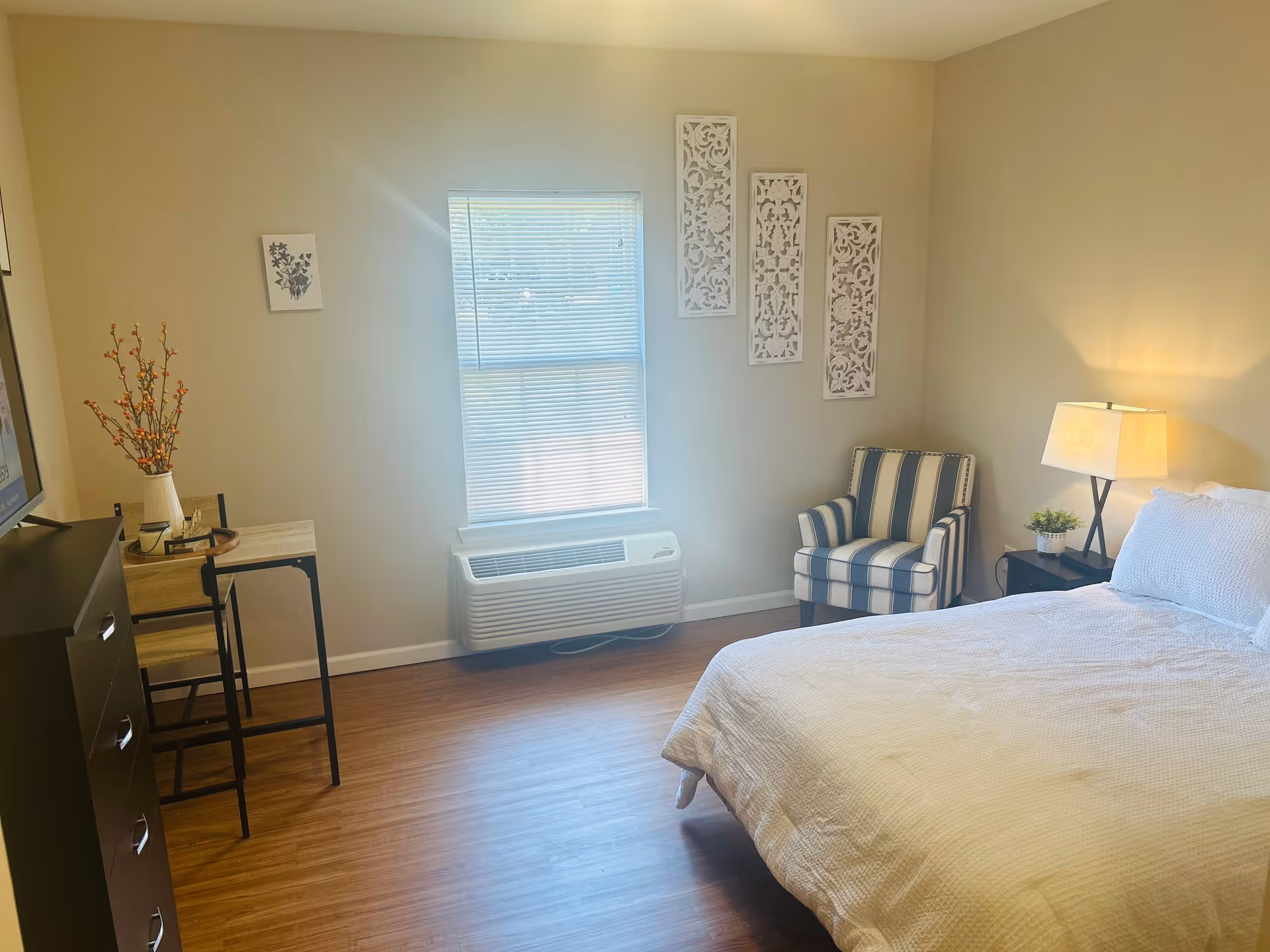 A bright bedroom with a large window covered by blinds, a bed with white bedding, a striped armchair, a nightstand with a lamp and a small plant, a tall dresser, and a small table with two chairs. The walls are decorated with three white carved panels and a small framed picture.
