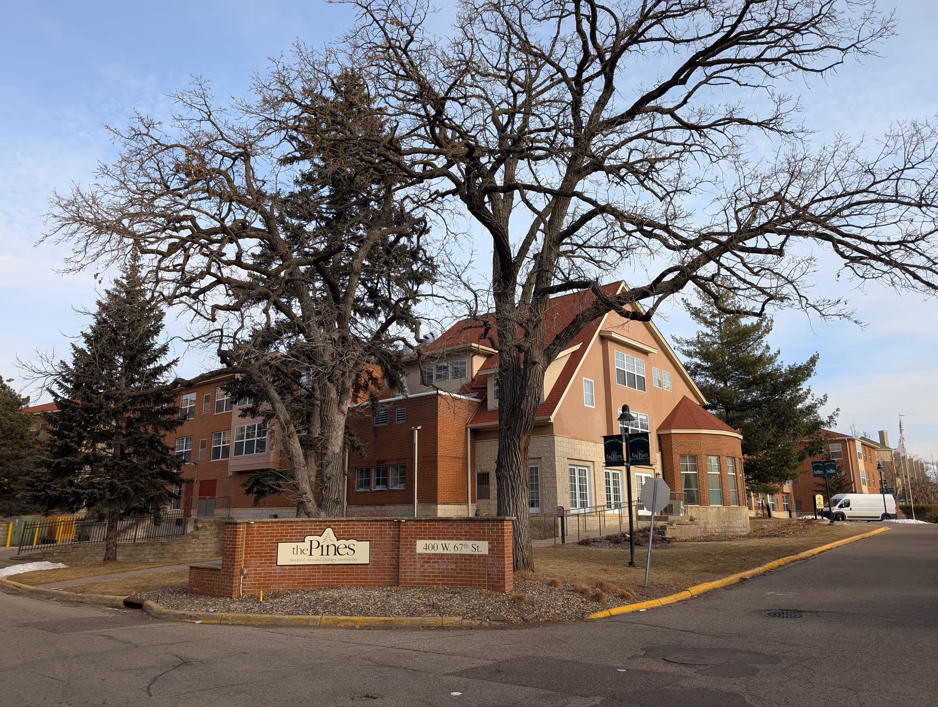 Exterior view of The Pines Senior and Assisted Living facility showing a multi-story building with a mix of brick and stone facade, large windows, and a red roof. There are leafless trees and evergreen trees in front of the building, a brick sign with the facility name and address, and a street with a sidewalk.