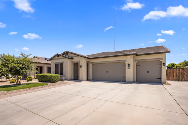 Exterior view of a single-story residential building with a three-car garage, beige stucco walls, and a tiled roof under a clear blue sky. There is a concrete driveway in front and some landscaping with bushes and a tree on the left side.