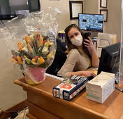 A woman wearing a face mask is sitting behind a reception desk with a computer monitor and a clear protective barrier. On the desk, there is a large wrapped bouquet of fruit, a box of gloves, and a box of face masks. Behind her, a monitor displays multiple security camera feeds, and framed certificates are visible on the wall.