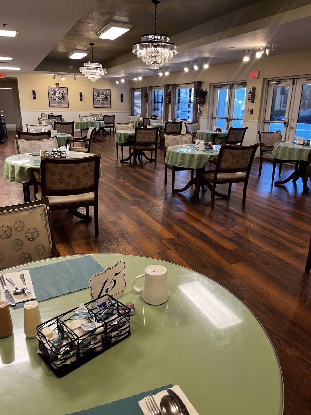 Dining room with round tables covered in green tablecloths, upholstered chairs, chandeliers, and hardwood floors.
