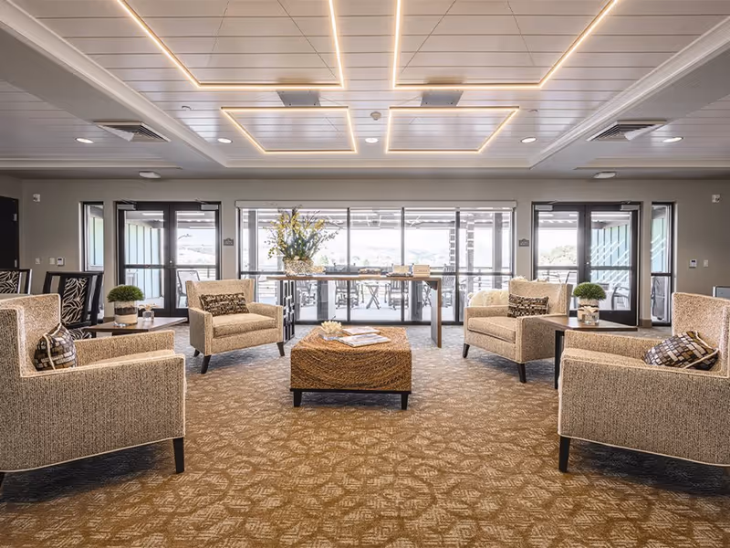 Bright, spacious communal lounge with armchairs arranged around a woven ottoman and large windows overlooking a patio.