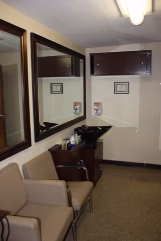 Interior of a small room with beige walls and carpeted floor, featuring two beige chairs, a large mirror on the wall, a black sink with hair care products on a small cabinet, and a wall-mounted cabinet above the sink.
