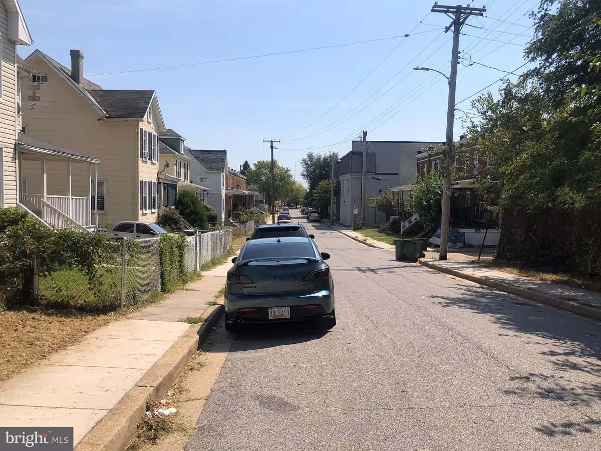 A residential street with parked cars along the curb, houses with porches and fenced yards on the left side, and trees and more houses on the right side under a clear blue sky.