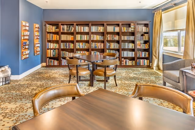 Comfortable reading room with bookshelves along a blue wall, round tables and leather chairs near a window with curtains.