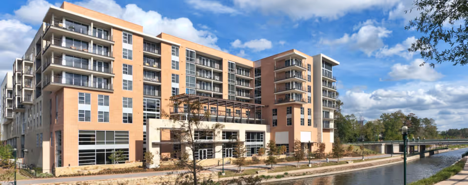 Exterior view of a multi-story senior living facility named Village at Woodlands Waterway, situated next to a waterway with a pedestrian bridge in the background under a partly cloudy sky.