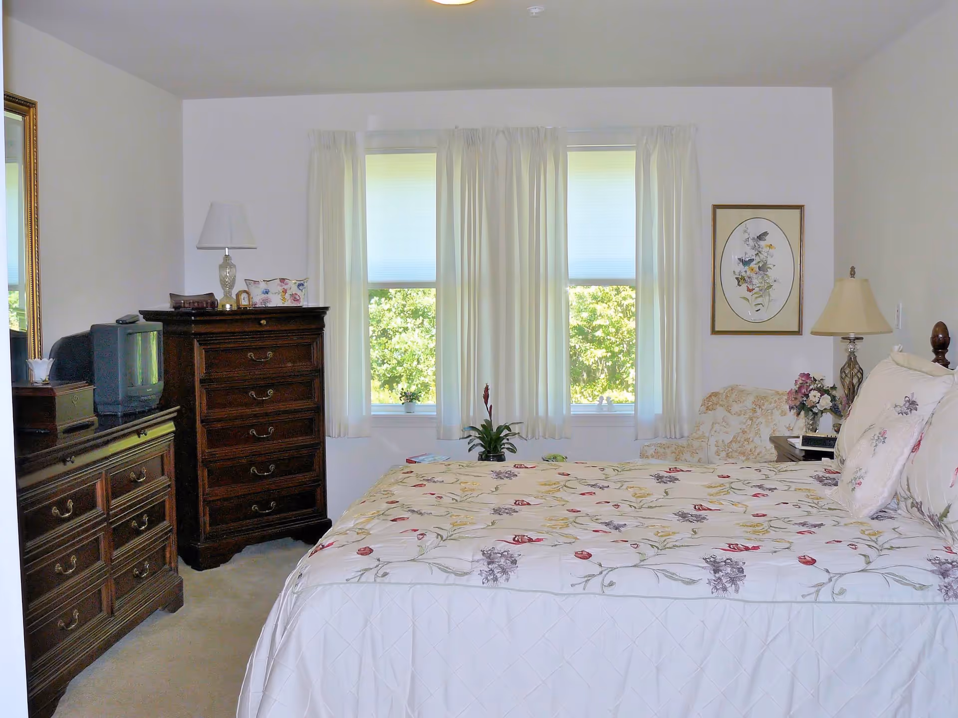 A bright bedroom with a large bed covered in a floral patterned bedspread and matching pillows. There are two windows with white curtains letting in natural light. The room features dark wooden furniture including a dresser with a mirror and a tall chest of drawers. A small TV sits on the dresser. There is a floral armchair next to a nightstand with a lamp and a flower arrangement. A framed floral artwork hangs on the wall.