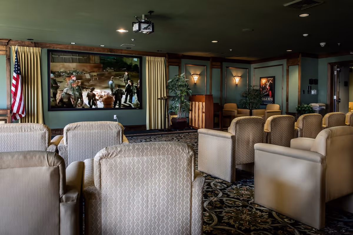 A cozy movie theater room with multiple beige upholstered armchairs arranged in rows facing a large screen showing a movie scene. The room has green walls, decorative wall sconces, an American flag, potted plants, and a piano in the corner.