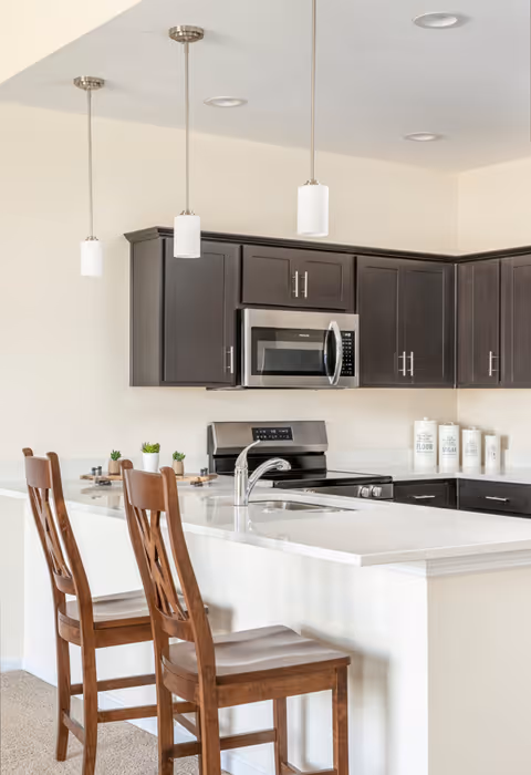 Modern kitchen with dark wood cabinets, a stainless steel microwave and stove, a white countertop island with a built-in sink, and two wooden bar stools. Three pendant lights hang above the island, and there are decorative containers labeled flour, sugar, coffee, and tea on the countertop.