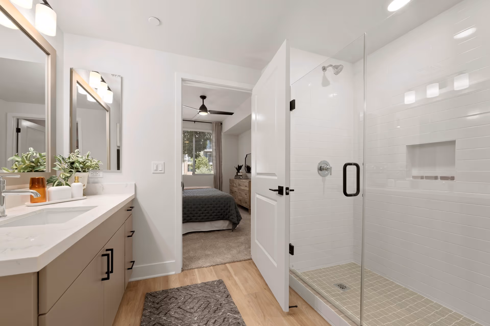 Modern bathroom with a double-sink vanity, glass-enclosed shower, and an open door leading into a bedroom.