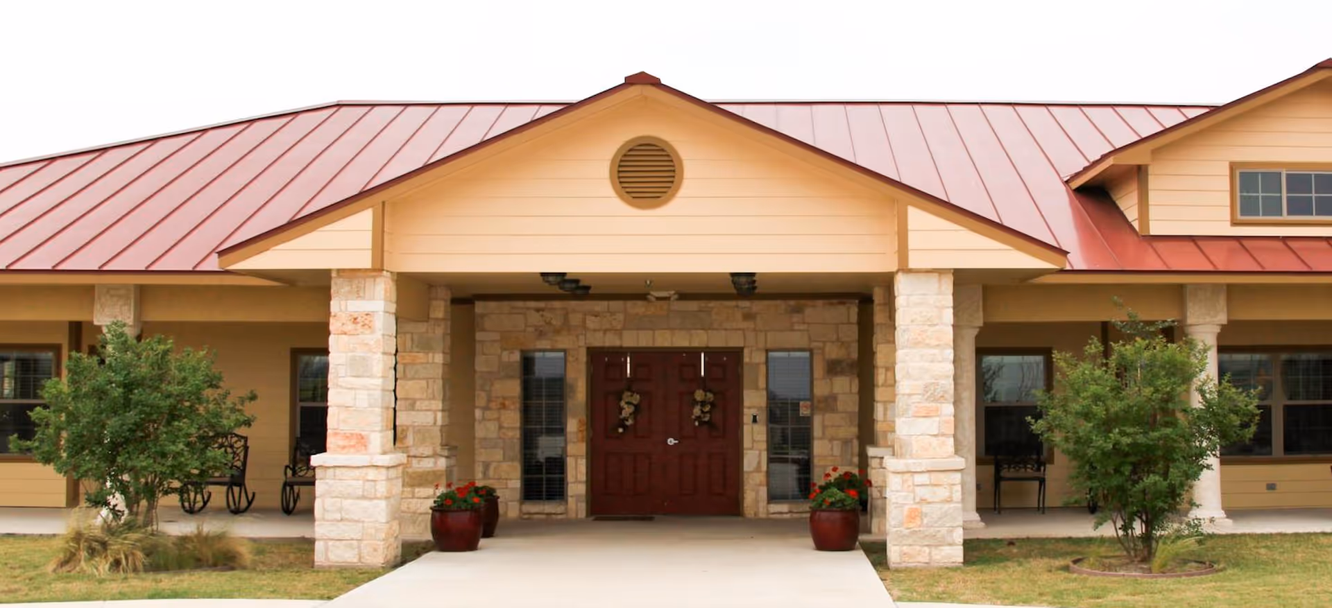 Front entrance of a single-story senior living building with stone columns, a covered portico and red double doors under a red metal roof.