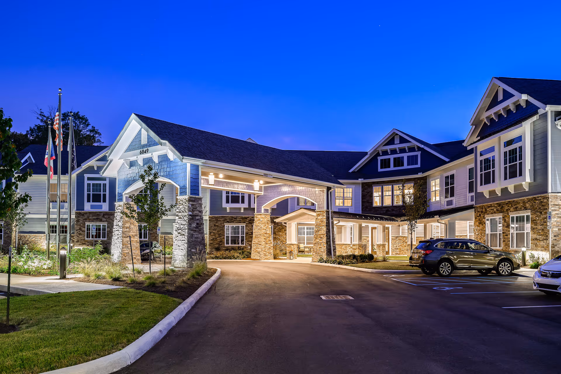 Exterior view of The Ashton at Anderson senior living facility at dusk, showing a well-lit entrance with stone pillars, multiple windows, and parked cars in front.