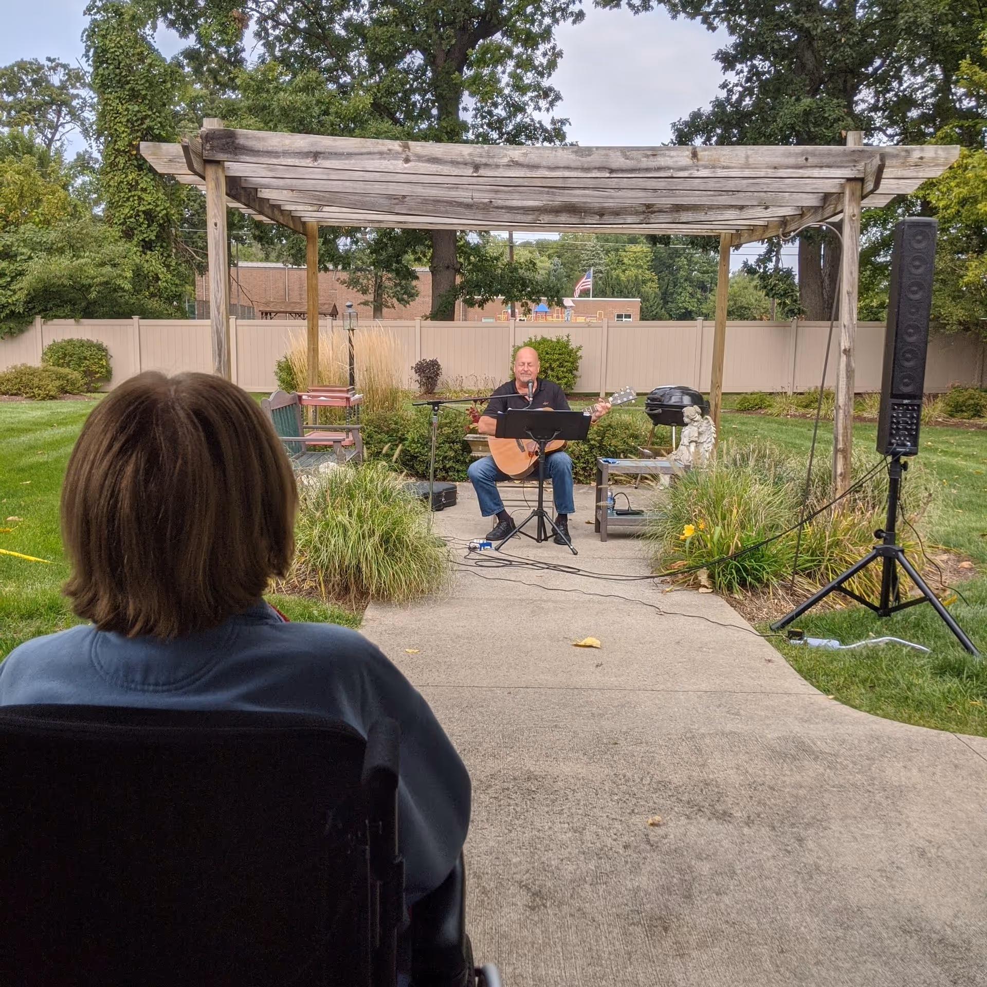 An elderly person in a wheelchair watches a man playing guitar and singing under a wooden pergola in an outdoor garden area. The setting includes green grass, bushes, and trees, with a beige fence and a building in the background.