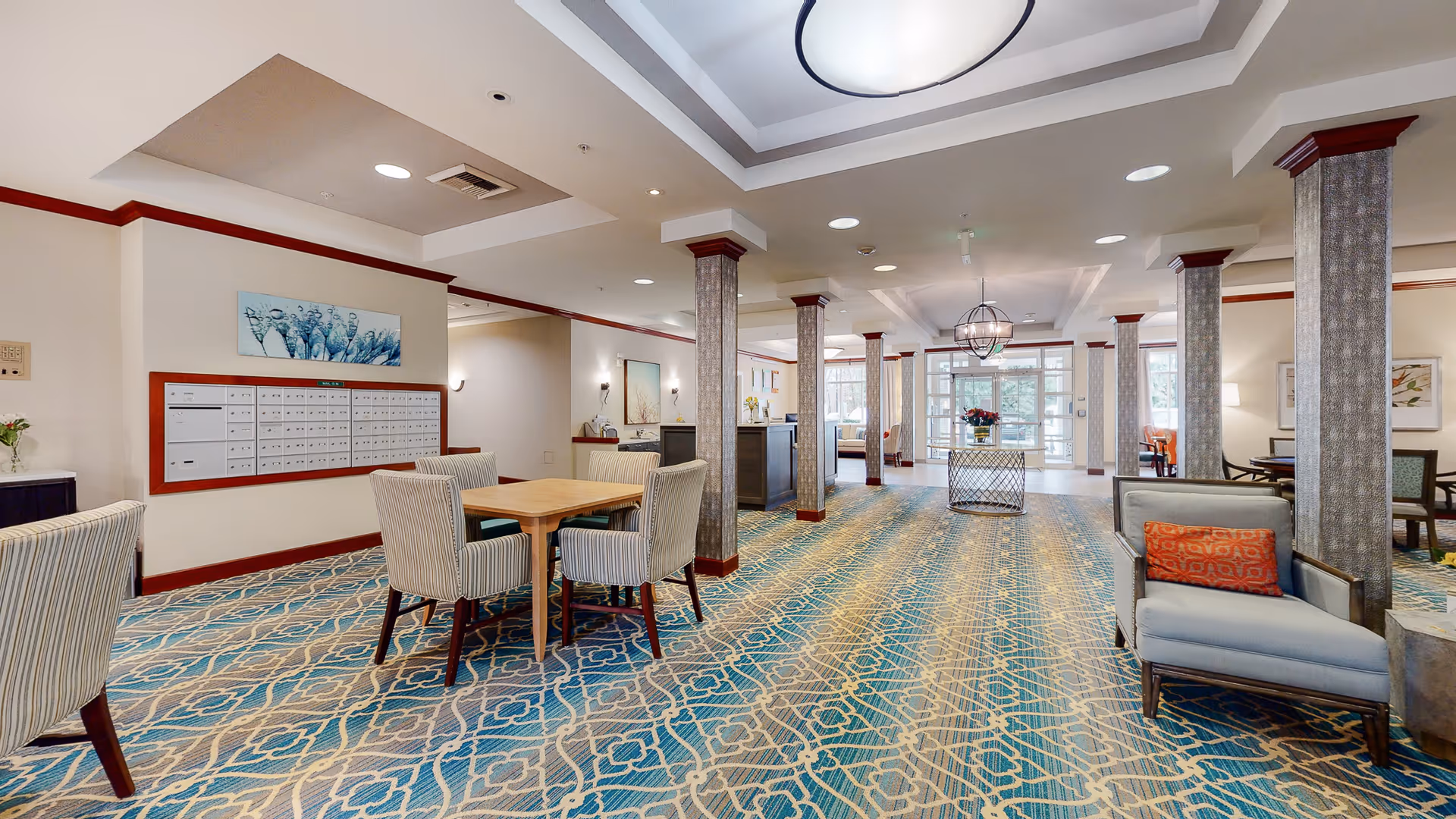 Spacious interior common area with patterned carpet, columns, seating and a table near a wall of mailboxes.