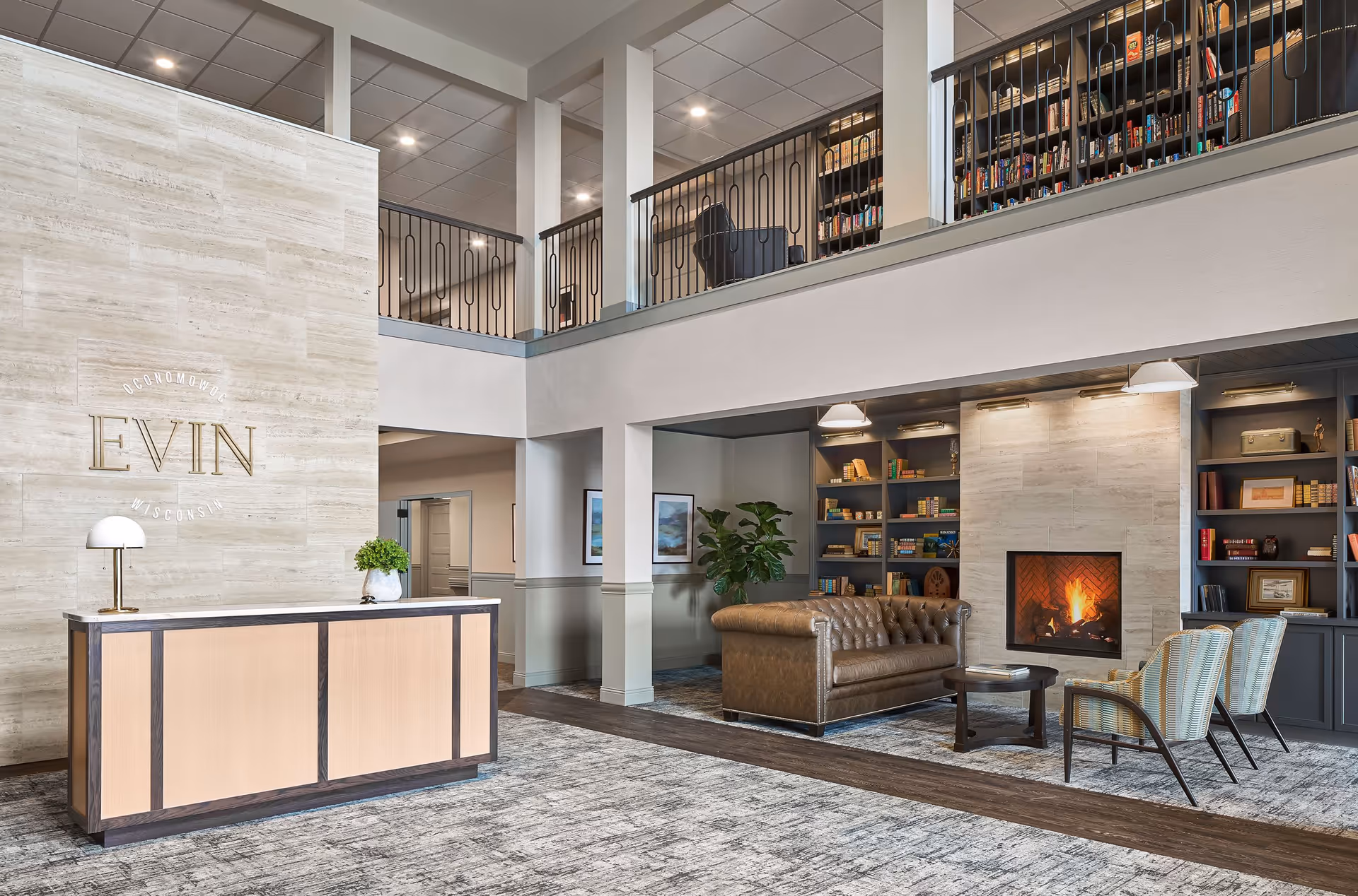 Interior view of a senior living facility lobby with a reception desk on the left and a cozy seating area on the right featuring a brown leather sofa, two armchairs, a coffee table, built-in bookshelves, and a lit fireplace. The space has a high ceiling with a mezzanine level and modern lighting.