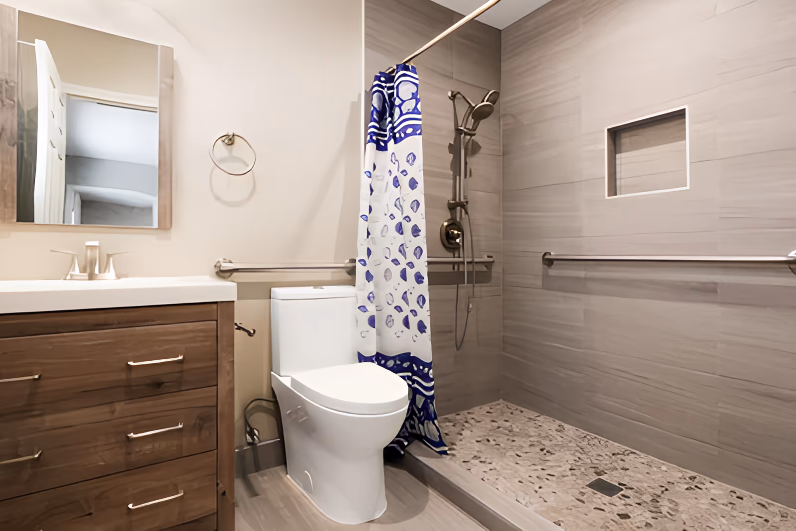 A modern bathroom featuring a wooden vanity with a white sink and silver faucet, a rectangular mirror above it, a white toilet, and a walk-in shower with a blue and white patterned shower curtain. The shower area has beige tiled walls, a built-in niche, and a handheld showerhead with grab bars installed for safety.