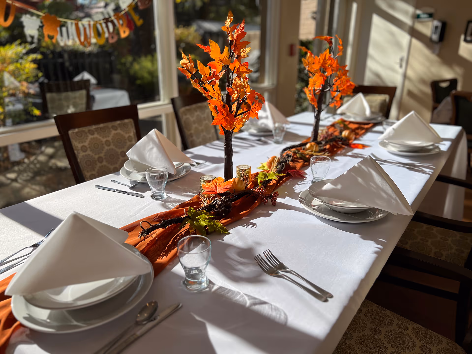 Sunlit dining table set with white linens, folded napkins, glassware, and orange autumn leaf centerpieces.