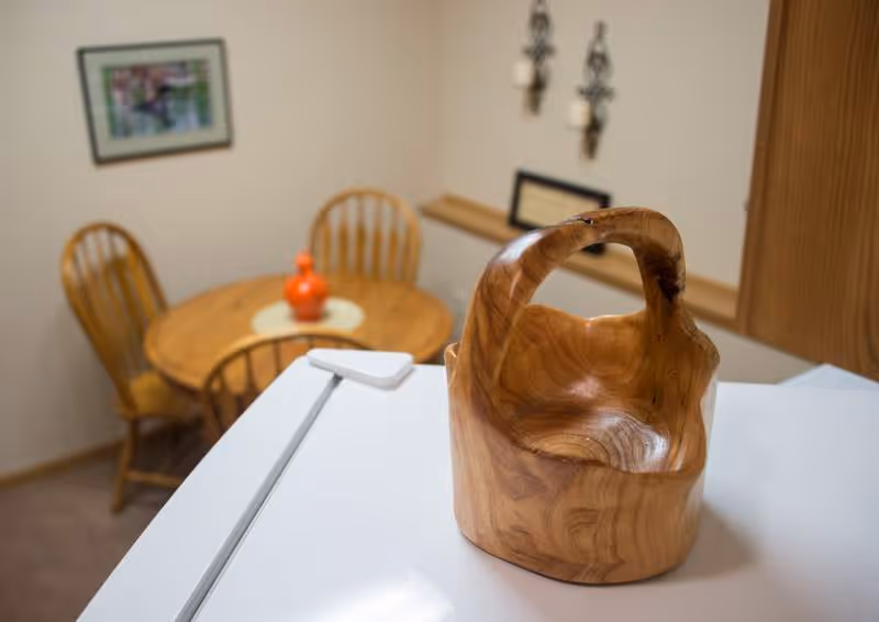 A wooden decorative basket placed on a white surface with a small round wooden dining table and four wooden chairs in the background. The room has beige walls with framed pictures and wall-mounted candle holders.