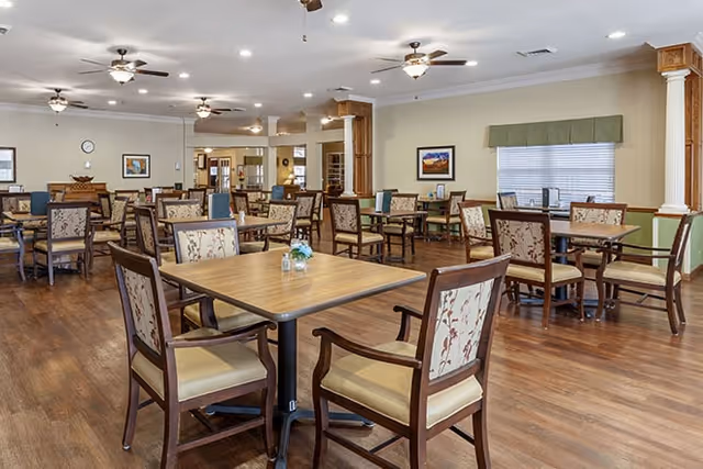 A spacious dining room with multiple wooden tables and cushioned chairs arranged neatly on a polished wooden floor. The room is well-lit with ceiling lights and fans, and decorated with framed pictures on the walls and a window with a green valance.