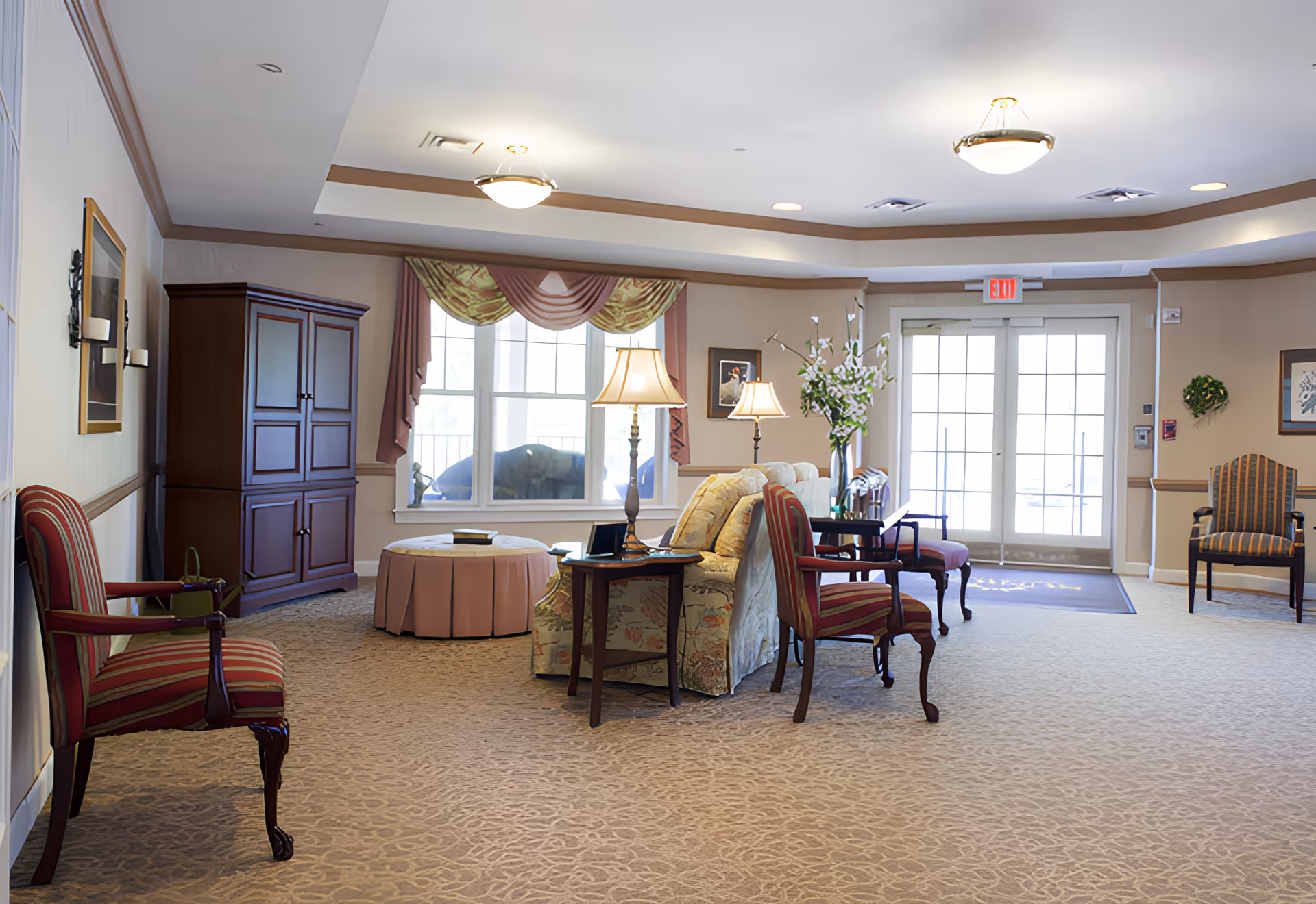 A spacious and well-lit living room area in a senior living facility with patterned carpet, multiple upholstered chairs, a floral sofa, side tables with lamps, a round ottoman, large windows with drapes, and double glass doors leading outside. The room has beige walls with wooden trim and framed artwork.