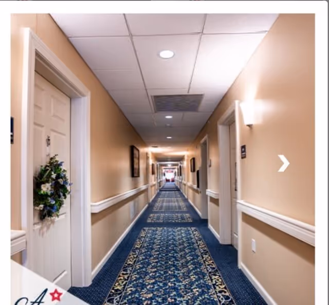 A long, well-lit hallway in an assisted living facility with beige walls, white doors on both sides, and a blue patterned carpet runner. One door has a decorative wreath. The ceiling has recessed lighting and ventilation panels.