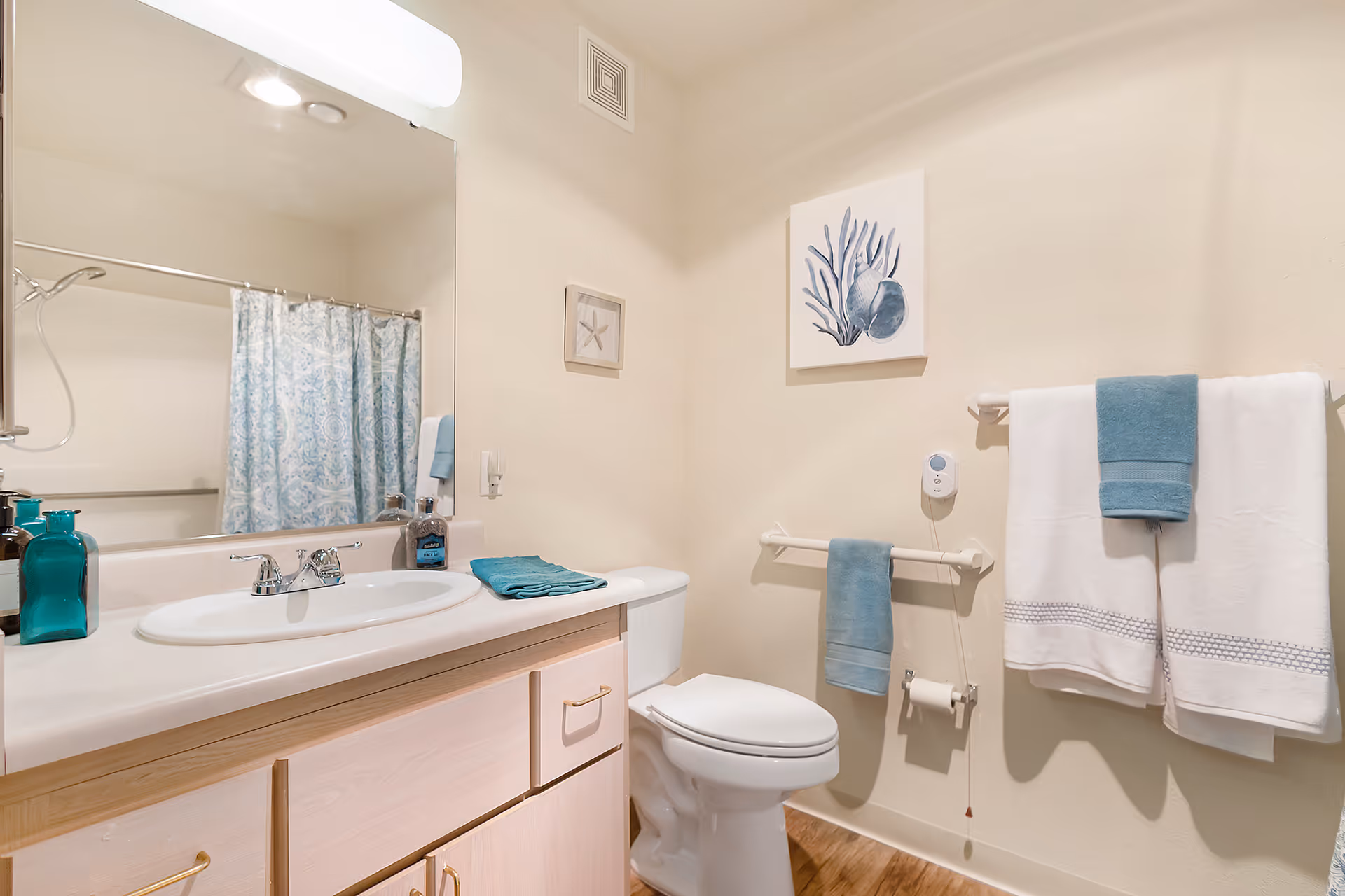 A clean and well-lit bathroom featuring a white sink with a large mirror above it, a toilet, and a shower with a blue patterned curtain. The bathroom has light-colored walls and wooden cabinetry under the sink. Blue and white towels hang on towel racks, and there are decorative wall art pieces with sea-themed designs.