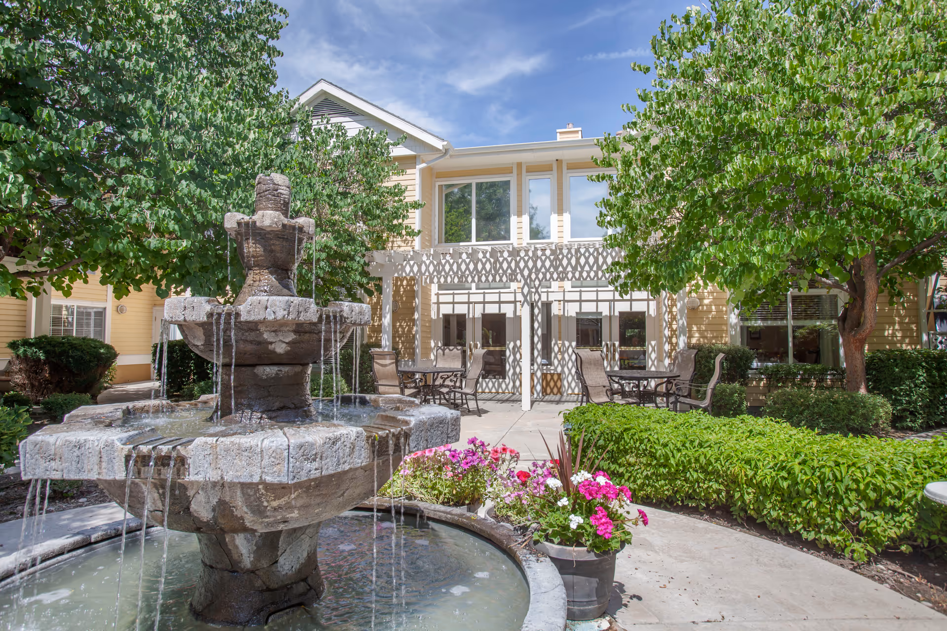 Stone tiered fountain in a landscaped courtyard with patio tables and a two-story building in the background.