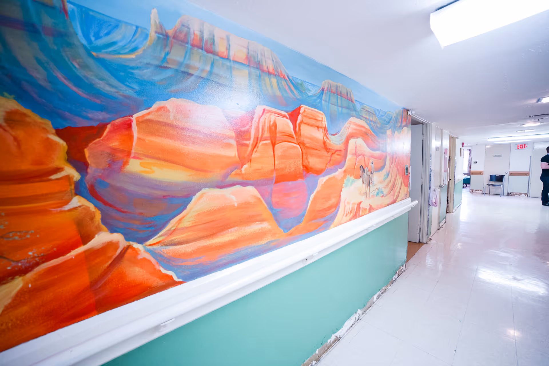 A brightly lit hallway with a colorful mural depicting a desert landscape with red rock formations and a person riding a horse. The hallway has light green lower walls and white upper walls and floor. There are doors and chairs along the hallway, and a person is standing near the end of the corridor.