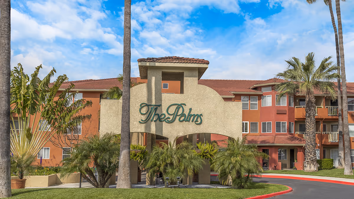 Exterior view of a senior living facility named The Palms, featuring a large beige entrance sign with green lettering, surrounded by palm trees and greenery, with a multi-story building with red and orange walls and tiled roof in the background under a partly cloudy blue sky.