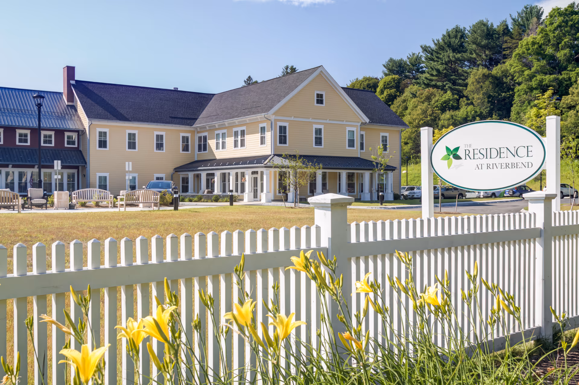 Exterior view of The Residence at Riverbend, a two-story yellow building with white trim, surrounded by a white picket fence and yellow flowers in the foreground under a clear blue sky.