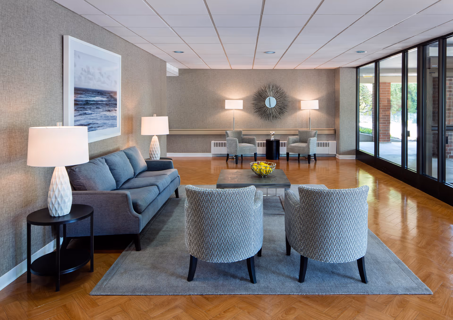 A modern and spacious living room area with a gray sofa, two patterned armchairs, a coffee table with a bowl of lemons, two side tables with lamps, and a large window wall letting in natural light. The room features a decorative wall mirror and two additional armchairs with floor lamps in the background.