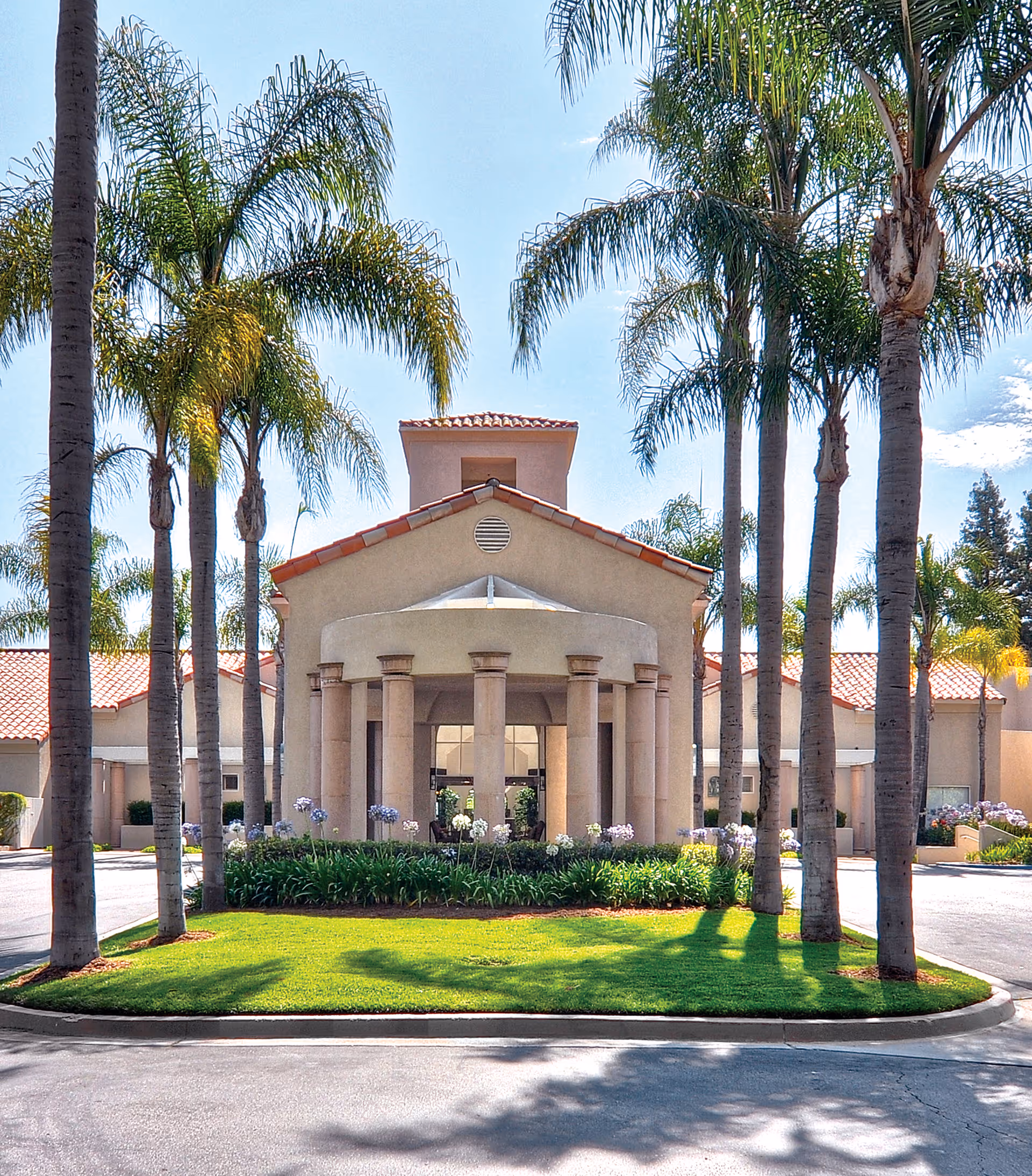 Front entrance of a senior living building with columns, a red tile roof, palm trees, and a circular driveway.