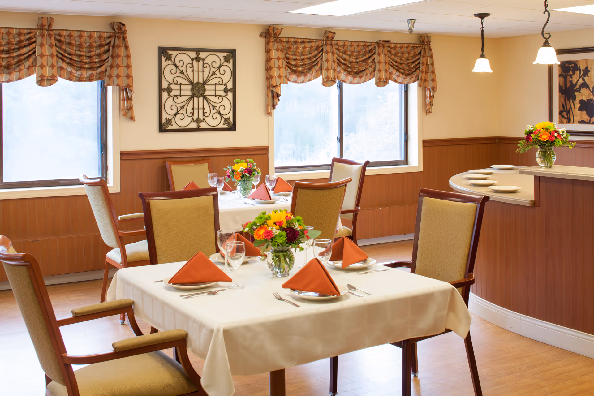 A dining room with tables set for a meal, featuring white tablecloths, orange folded napkins, glassware, and floral centerpieces. The room has wooden paneling on the walls, patterned valances on the windows, and pendant lights hanging from the ceiling.