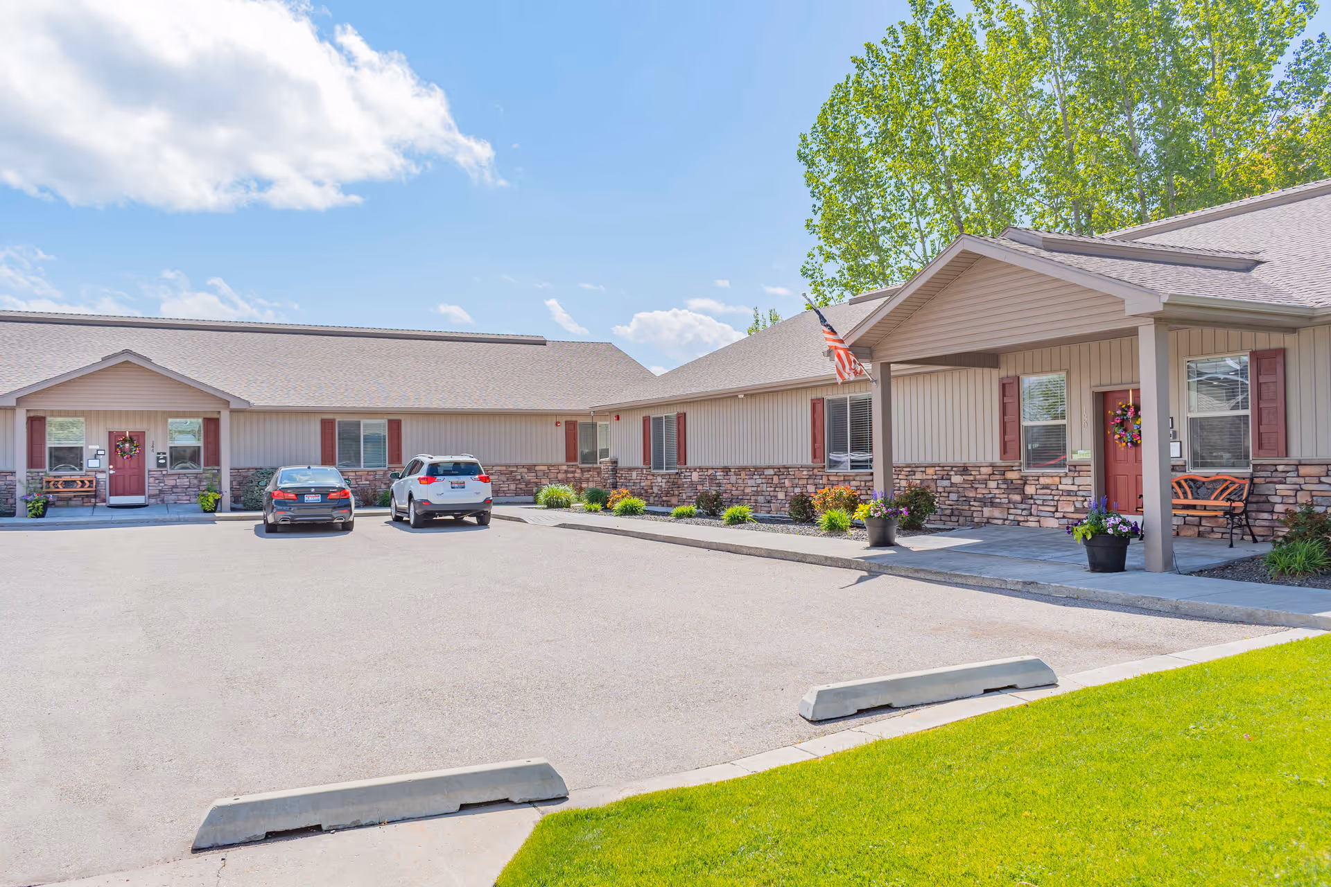 Exterior front of a single-story senior living building with a parking lot, two cars, and an American flag.