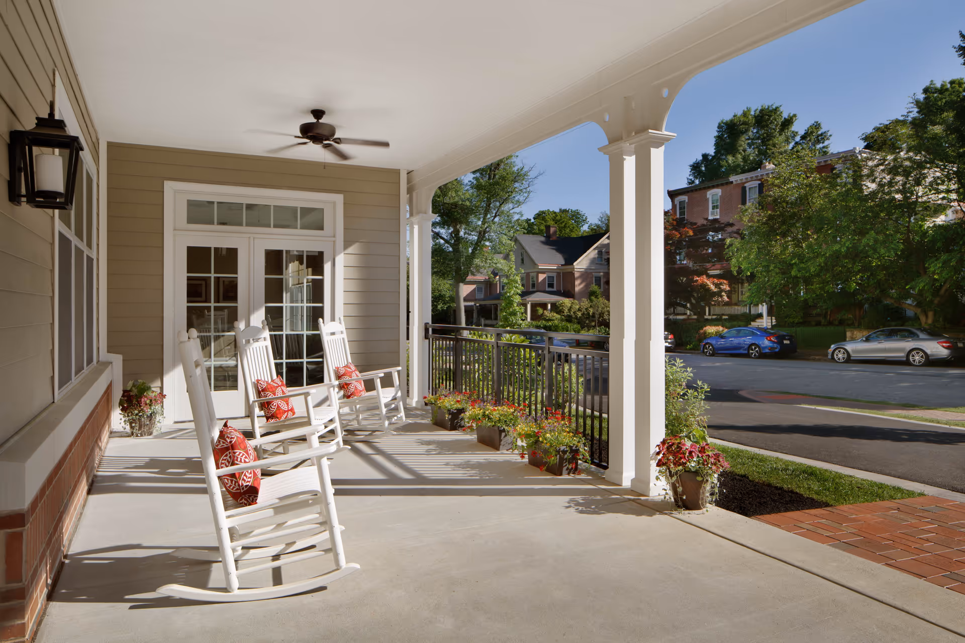 A covered porch area with three white rocking chairs, each with a red patterned cushion. The porch has a ceiling fan, potted plants along the railing, and a view of a quiet street with parked cars and houses in the background.
