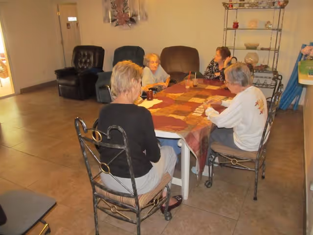 Four elderly women sitting around a rectangular table covered with a patchwork tablecloth in a living room area. The room has tiled floors, several cushioned chairs, and a metal shelving unit with decorative items. The women appear to be engaged in a card game or similar activity.