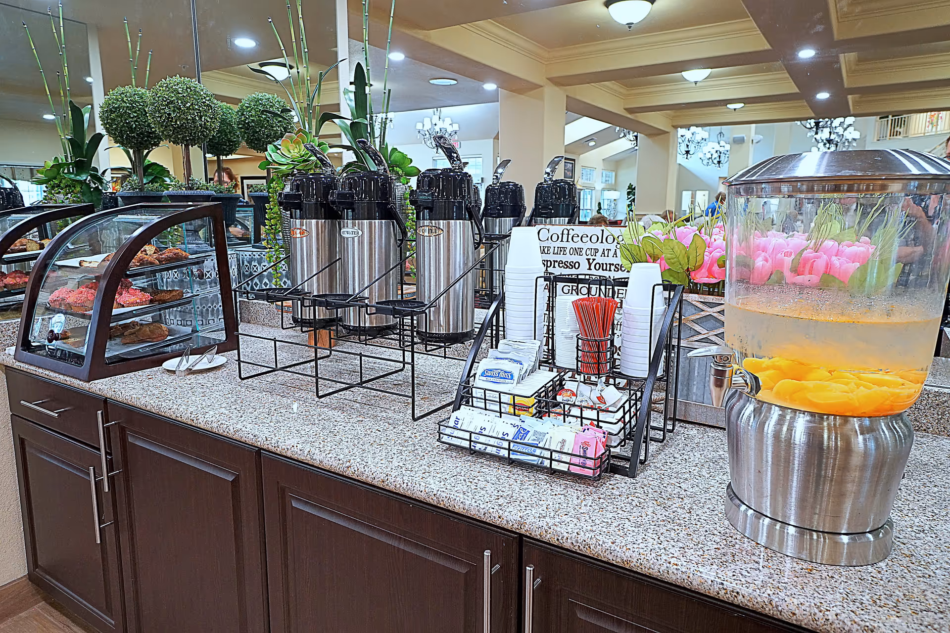 A beverage and snack station in a senior living facility featuring a granite countertop with multiple stainless steel coffee dispensers, a glass container with peach-infused water, a display case with pastries, and a rack holding disposable cups, sweeteners, and stirrers. The background shows a well-lit interior with plants and mirrors.