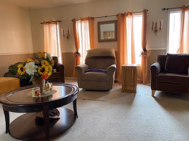 A cozy living room with beige carpet, two brown armchairs, and a brown recliner chair in the center. There are three windows with peach-colored curtains letting in natural light. A wooden coffee table with a glass top holds a vase of colorful flowers and two small decorative cups. A small wooden side table is next to the recliner, and a mirror hangs on the wall between the windows.