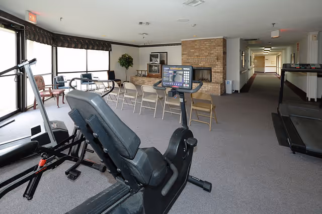 Interior room with exercise equipment including a recumbent bike and treadmill, several folding chairs arranged in rows, a brick fireplace, a TV on a stand, a potted plant, and large windows letting in natural light.
