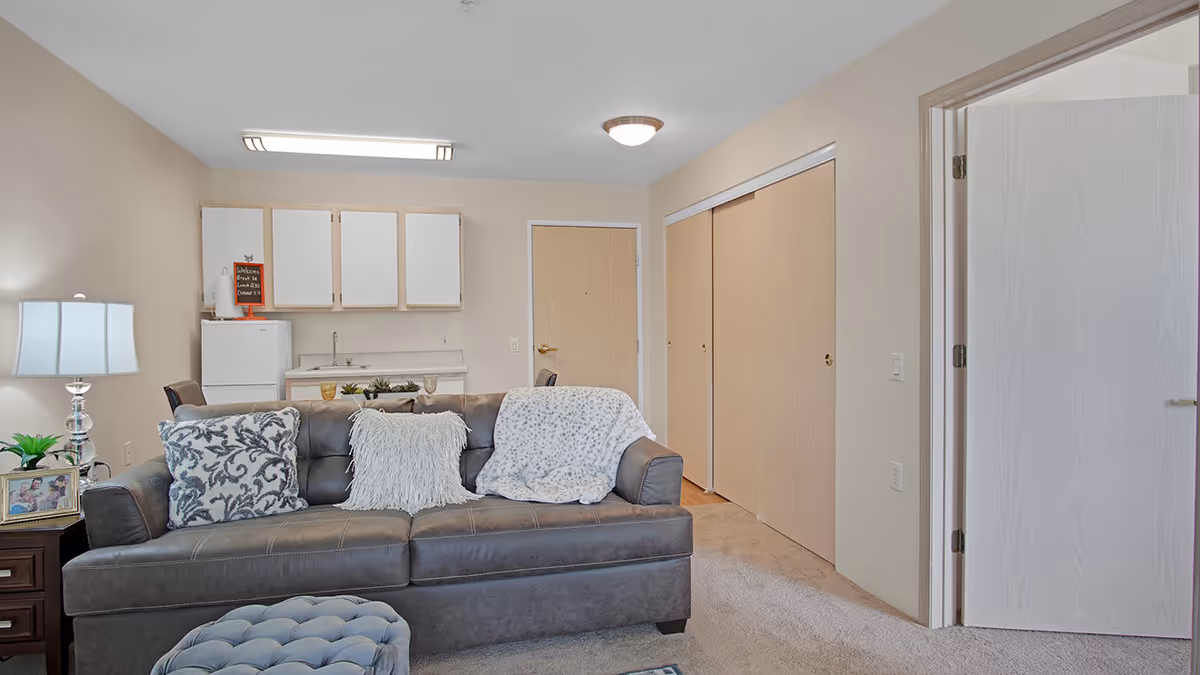 Interior view of a senior living facility unit at Holiday Grasslands Estates showing a living area with a gray sofa adorned with patterned and fluffy pillows, a small ottoman, a side table with a lamp and framed photo, and a kitchenette in the background with white cabinets, a small refrigerator, and a sink. There are two closed wooden doors and an open door leading to another room.