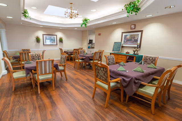 Dining room with several tables set with purple tablecloths and green napkins, wooden chairs, and hardwood flooring under soft ceiling lighting.
