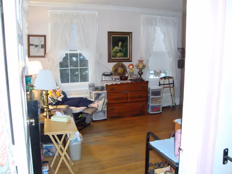 A cozy room with wooden floors and two windows with white curtains. There is a wooden dresser with decorative items and a framed painting above it. A person is reclining on a dark-colored chair near the left window. Various small furniture pieces, including a side table with a lamp and a walker, are also visible.