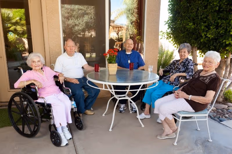 Five elderly individuals sitting around a round outdoor table with a potted plant in the center. Two women are seated on chairs, one woman is in a wheelchair, and two men are seated on chairs. They are outside near a building with large windows and surrounded by greenery.