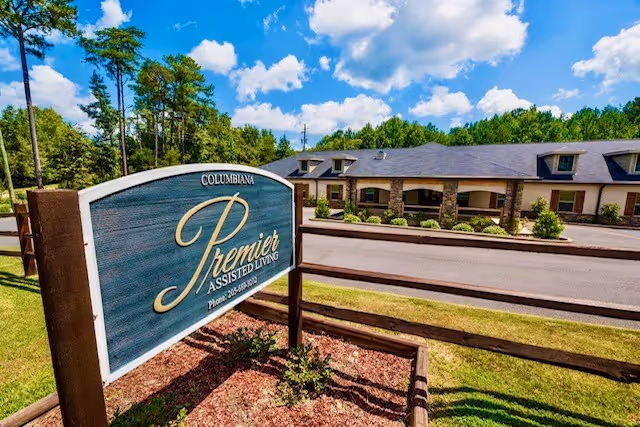 Outdoor view of Premier Assisted Living facility with a large sign in the foreground displaying the name and phone number. The building is single-story with a stone facade and surrounded by greenery and trees under a partly cloudy sky.