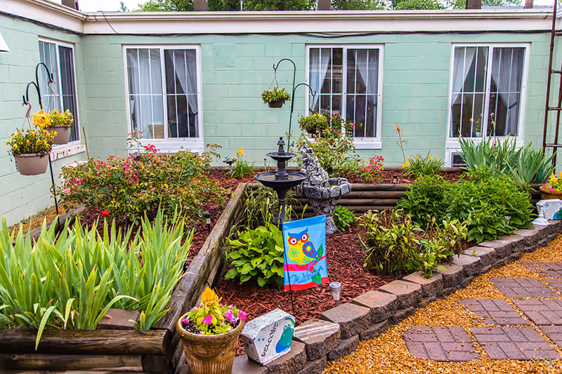 Small courtyard garden with raised flower beds, a birdbath fountain, decorative owl flag and hanging planters in front of pale green building windows.